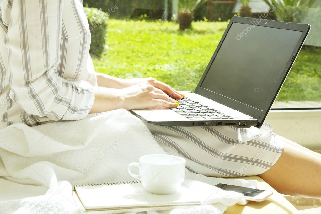 Beautiful hipster woman in loose stripe shirt sitting at home, using personal computer. Female writer writing screenplay at home, by big window, Drinking coffee. copy space, background, close up