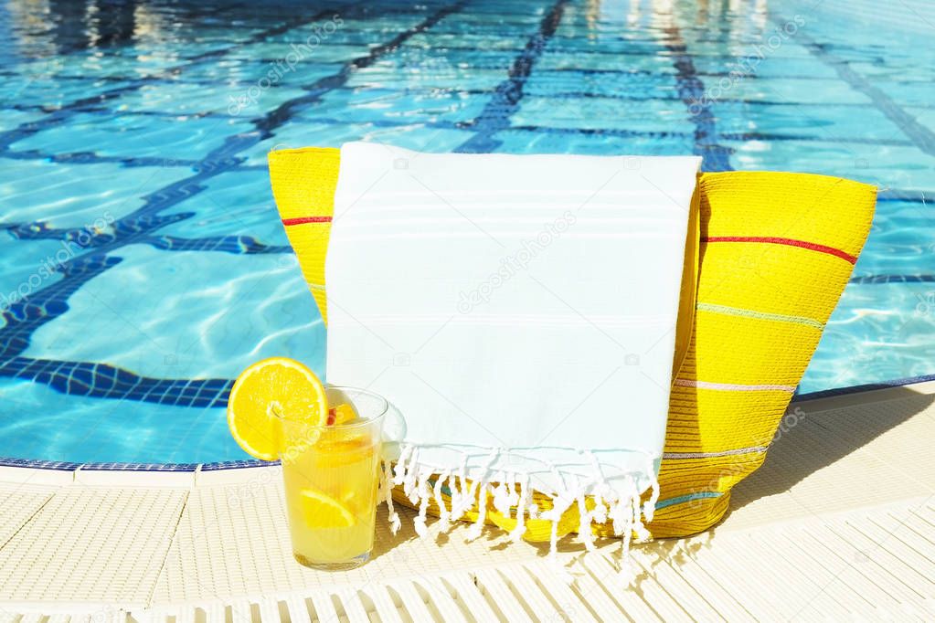 Poolside composition with orange, sunscreen lotion and beach / bath accessories. Beautiful clean big pool on sunny day,