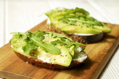 Vejetaryen sandviç, çavdar ekmeği tost, avokado, vegenaise eggless mayonez sos, baharat susam, roka rucola yaprakları Beyaz ahşap doku tablo. Ulusal avokado gün kavramı. Yakın çekim, arka plan