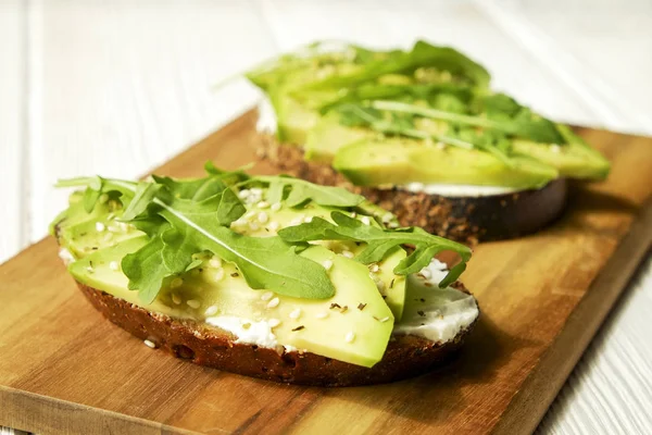 Vegan sandwich roggebrood toast, avocado, vegenaise eggless mayonaise saus, sesamzaad, kruiden, rucola rucola bladeren wit houtstructuur tabel. Nationale dag van de avocado concept. Close-up, achtergrond — Stockfoto