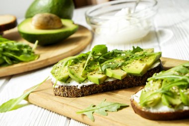 Vejetaryen sandviç, çavdar ekmeği tost, avokado dilimleri, vegenaise sos ve ham roka. Kızarmış hamur mayası, yumurtasız mayonez, ahşap masa, bütün ve yarıya. Ulusal avokado gün kavramı. Yakın çekim, arka plan.