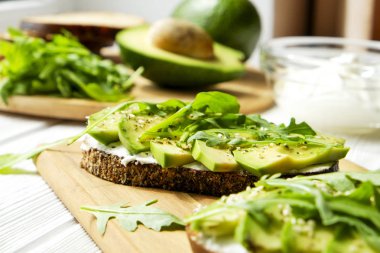 Vejetaryen sandviç, çavdar ekmeği tost, avokado dilimleri, vegenaise sos ve ham roka. Kızarmış hamur mayası, yumurtasız mayonez, ahşap masa, bütün ve yarıya. Ulusal avokado gün kavramı. Yakın çekim, arka plan.