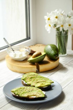 Vejetaryen sandviç, çavdar ekmeği tost, avokado, bütün ve yarıya, vegenaise sos. Kızarmış hamur mayası, yumurtasız mayonez, ahşap dilim, nergis çiçek vazo. Ulusal avokado gün kavramı. Yakın çekim, arka plan.