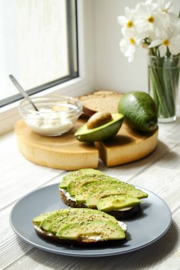 Vejetaryen sandviç, çavdar ekmeği tost, avokado, bütün ve yarıya, vegenaise sos. Kızarmış hamur mayası, yumurtasız mayonez, ahşap dilim, nergis çiçek vazo. Ulusal avokado gün kavramı. Yakın çekim, arka plan.