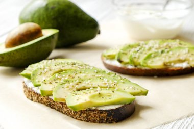 Vejetaryen sandviç, çavdar ekmeği tost, avokado dilimleri, vegenaise sos ve ham roka. Kızarmış hamur mayası, yumurtasız mayonez, ahşap masa, bütün ve yarıya. Ulusal avokado gün kavramı. Yakın çekim, arka plan.