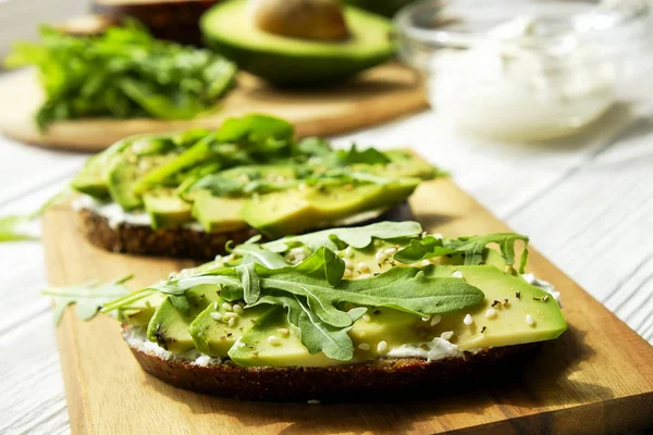 Vegan sandwich, roggebrood toast, avocado segmenten, vegenaise saus & rauwe rucola. Geroosterde zuurdesem, eggless mayonaise, houten tafel, hele & gehalveerde. Nationale dag van de avocado concept. Close-up, achtergrond. — Stockfoto