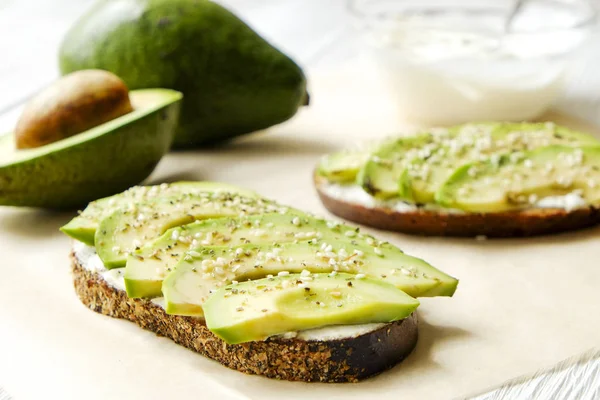Vejetaryen sandviç, çavdar ekmeği tost, avokado dilimleri, vegenaise sos ve ham roka. Kızarmış hamur mayası, yumurtasız mayonez, ahşap masa, bütün ve yarıya. Ulusal avokado gün kavramı. Yakın çekim, arka plan.
