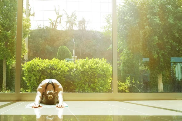 Yogi woman does yoga exercises, working out, practicing pranayama breathing technique. Spacious room with big full length windows.