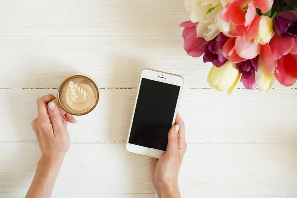 Overhead shot of woman hands holding cell phone gadget & coffee cup w cappuccino latte art on white wooden table background. Beautiful tulip flowers bouquet of different colors. Copy space, close up.