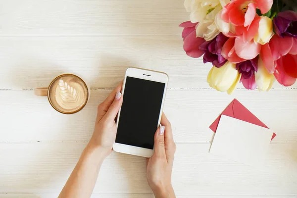Kadın el cep telefonu gadget ve kahve holding genel gider atış Kupası w cappuccino latte sanat Beyaz ahşap masa arka plan üzerinde. Farklı renklerde güzel Lale çiçek buketi. Uzay kopyalayın, yakın çekim.