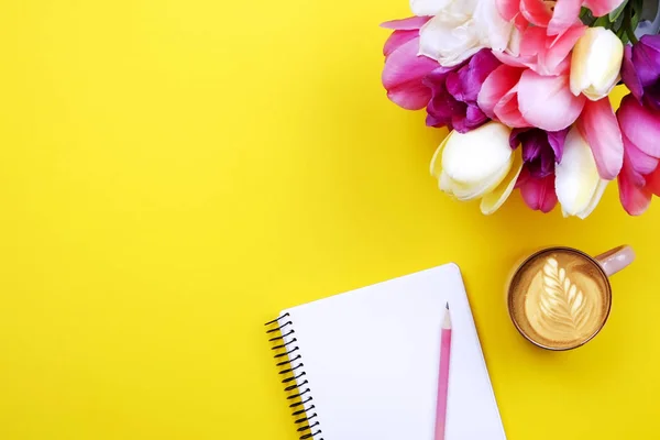 Composição de mesa feminina com caderno de folha em branco, lápis, xícara de café, buquê de tulipas no fundo amarelo. Local de trabalho feminino, flores para o dia da mãe. Vista superior, flat lay, close up, espaço de cópia . — Fotografia de Stock