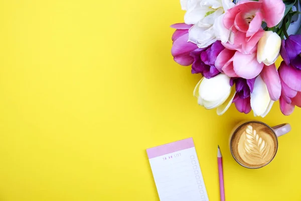 Composição de mesa feminina com caderno de folha em branco, lápis, xícara de café, buquê de tulipas no fundo amarelo. Local de trabalho feminino, flores para o dia da mãe. Vista superior, flat lay, close up, espaço de cópia . — Fotografia de Stock