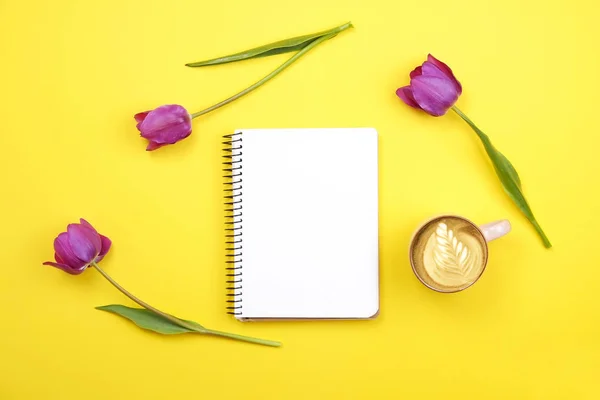 Composição do desktop feminino com caderno de folha em branco, café xícara de arte latte, buquê de tulipas no fundo amarelo. Local de trabalho feminino, flores para o dia da mãe. Vista superior, flat lay, close up, espaço de cópia . — Fotografia de Stock