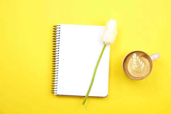 Composição do desktop feminino com caderno de folha em branco, café xícara de arte latte, buquê de tulipas no fundo amarelo. Local de trabalho feminino, flores para o dia da mãe. Vista superior, flat lay, close up, espaço de cópia . — Fotografia de Stock