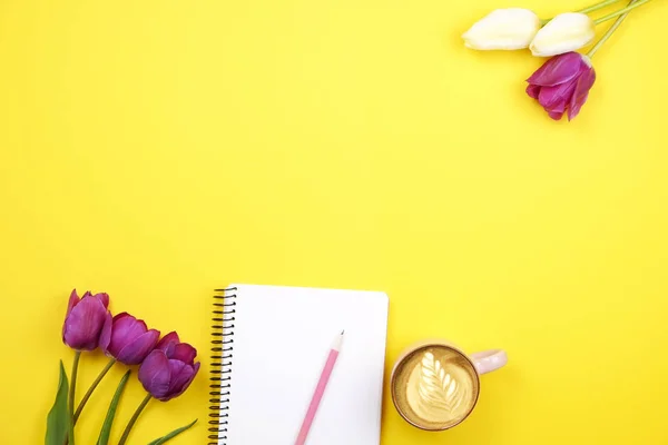 Composição de mesa feminina com caderno de folha em branco, lápis, xícara de café, buquê de tulipas no fundo amarelo. Local de trabalho feminino, flores para o dia da mãe. Vista superior, flat lay, close up, espaço de cópia . — Fotografia de Stock