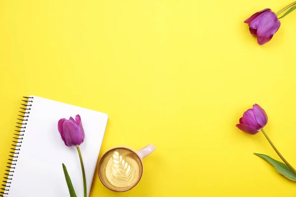 Feminine desktop composition with blank sheet notebook, coffee cup latte art, tulips bouquet on yellow background. Girlie workplace, flowers for mother\'s day. Top view, flat lay, close up, copy space.