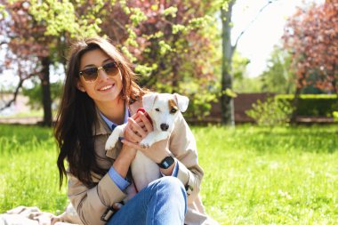 Portrait of attractive young woman hugging cute jack russell terrier puppy in park, green lawn, foliage background. Hipster female in sunglasses smiling, pets her dog on grass. Close up, copy space. clipart