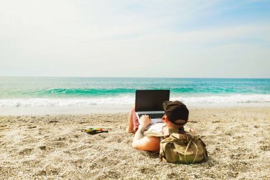 Genç adam atlet, şort, dizüstü bilgisayar ve sırt çantası ile plajda snapback kap. Serbest meslek, hipster blogger, deniz manzarayı yazar