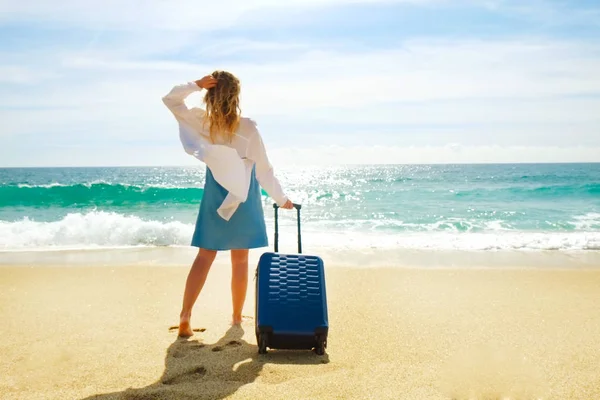 Hipster young woman on vacation in denim dress, white cotton shirt ombre hair walking to sea with suitcase on perfect sunny day.