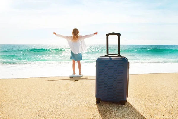 Hipster young woman on vacation in denim dress, white cotton shirt ombre hair walking to sea with suitcase on perfect sunny day.