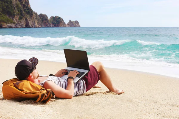 Joven hombre en camiseta sin mangas, pantalones cortos, gorra snapback en la playa con portátil y mochila. Freelancer, blogger hipster, escritor disfrutando de la vista al mar —  Fotos de Stock