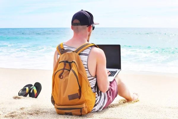 Joven hombre en camiseta sin mangas, pantalones cortos, gorra snapback en la playa con portátil y mochila. Freelancer, blogger hipster, escritor disfrutando de la vista al mar Imagen De Stock
