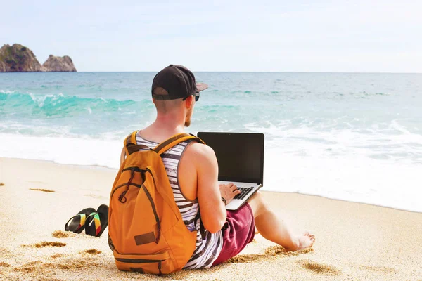 Joven hombre en camiseta sin mangas, pantalones cortos, gorra snapback en la playa con portátil y mochila. Freelancer, blogger hipster, escritor disfrutando de la vista al mar Imagen De Stock