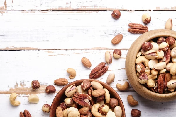 Trail mix with different kinds of nuts in brown wood bowl on scratched white wooden table background, soft daylight. Copy space.