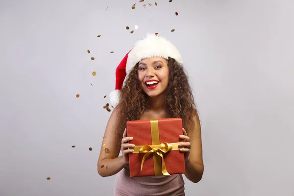 Studio portrait of young woman with dark skin and long curly hair wearing tight sexy dress, holding wrapped present with golden bow. Close up, copy space for text, isolated background. — Stock Photo, Image