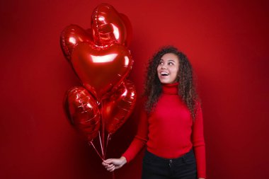 Kırmızı duvarın üzerinde poz veren muhteşem genç kadın elinde helyum şişirilmiş balon tutuyor..
