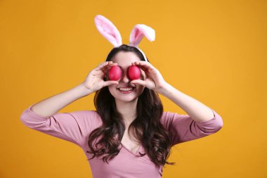 Paskalya için geleneksel tavşan kulaklığı takan ve gülümseyen genç bir kadının stüdyo portresi. Sarı arka planda dalgalı saçlı esmer kadın. Kapat, boşluğu kopyala.