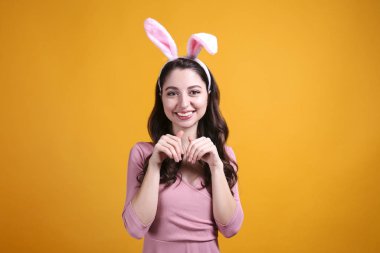 Paskalya için geleneksel tavşan kulaklığı takan ve gülümseyen genç bir kadının stüdyo portresi. Sarı arka planda dalgalı saçlı esmer kadın. Kapat, boşluğu kopyala.