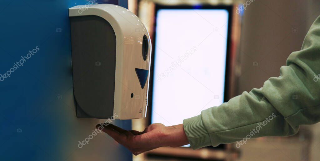 Macro shot of young woman using automatic hand sanitizer dispenser at the international airport with blank glowing advertisement banner on background. Close up, copy space.