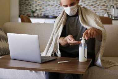 Koronavirüs karantina konsepti yüzünden evde çalışan yetişkin bir adamın yakın çekimi. Kanepede dizüstü bilgisayarla oturan erkek, önlem olarak dezenfektan kullanıyor. Arkaplan, boşluğu kopyala.