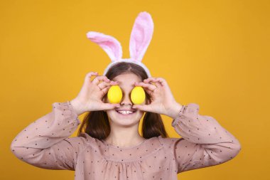 Paskalya 'da geleneksel tavşan kulaklığı takan genç bir kızın stüdyo portresi. Gözlerini yumurta ile kapatıp gülümsüyor. Sarı arka planda at kuyruklu esmer kadın. Kapat, boşluğu kopyala.