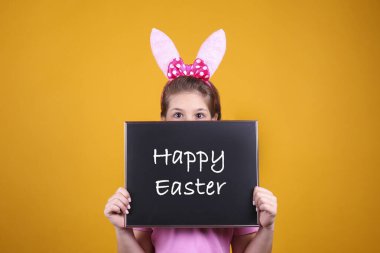 Geleneksel tavşan kulaklığı takan gülümseyen genç kızın stüdyo portresi. Saçları örgülü esmer kadın elinde mutlu paskalya metniyle karatahta tutuyor. Sarı arkaplan, kapat, boşluğu kopyala.