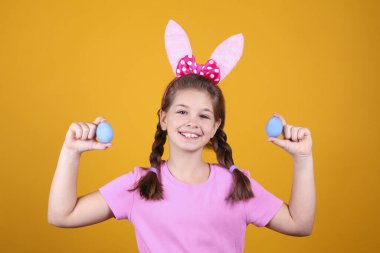 Paskalya 'da geleneksel tavşan kulaklığı takan gülümseyen genç kızın stüdyo portresi. Sarı arka planda geleneksel renkli yumurtaları tutan at kuyruklu esmer kadın. Kapat, boşluğu kopyala.