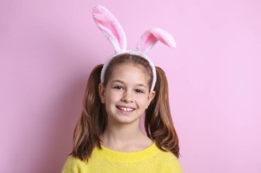Paskalya 'da geleneksel tavşan kulaklığı takan gülümseyen genç kızın stüdyo portresi. Saçları örgülü, açık sarı kazaklı esmer kadın portresi. Pembe arkaplan, kapat, boşluğu kopyala.
