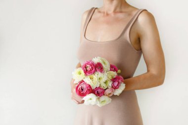 Elinde bahar çiçekleri tutan bej bir elbise giyen bir kadının kavramsal görüntüsü. Beyaz arka planda renkli beyaz ve pembe ranunculus buketi olan bir kadın. Kapat, kopyala boşluk, kırpılmış çekim.