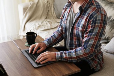 Kanepede oturan, dizüstü bilgisayar kullanan, internette gezinen, sıcak içecek içen tanınmamış bir adamın görüntüsü. Kendini soyutlama dönemi konsepti boyunca evden çalışmak. Kapat, kopyala boşluk, arkaplan.