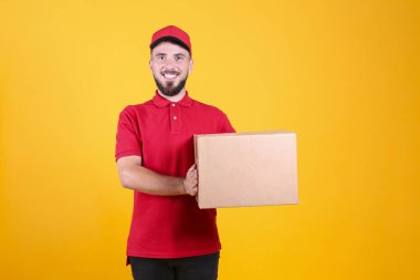 Kırmızı üniformalı ve şapkalı genç teslimatçı, boş karton kutuyu izole edilmiş beyaz arka planda tutuyor. Paket taşıyan dost canlısı sakallı adamın portresi. Metin için alanı kopyala.