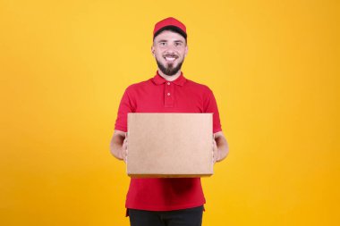 Kırmızı üniformalı ve şapkalı genç teslimatçı, boş karton kutuyu izole edilmiş beyaz arka planda tutuyor. Paket taşıyan dost canlısı sakallı adamın portresi. Metin için alanı kopyala.