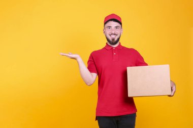 Kırmızı üniformalı ve şapkalı genç teslimatçı, boş karton kutuyu izole edilmiş beyaz arka planda tutuyor. Paket taşıyan dost canlısı sakallı adamın portresi. Metin için alanı kopyala.
