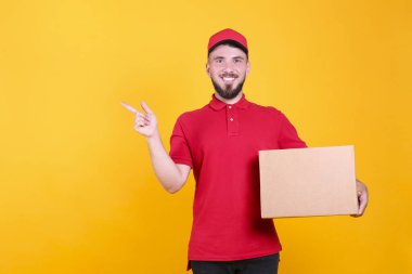 Kırmızı üniformalı ve şapkalı genç teslimatçı, boş karton kutuyu izole edilmiş beyaz arka planda tutuyor. Paket taşıyan dost canlısı sakallı adamın portresi. Metin için alanı kopyala.