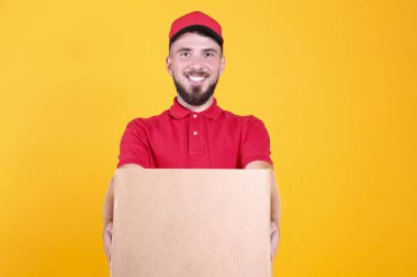 Kırmızı üniformalı ve şapkalı genç teslimatçı, boş karton kutuyu izole edilmiş beyaz arka planda tutuyor. Paket taşıyan dost canlısı sakallı adamın portresi. Metin için alanı kopyala.