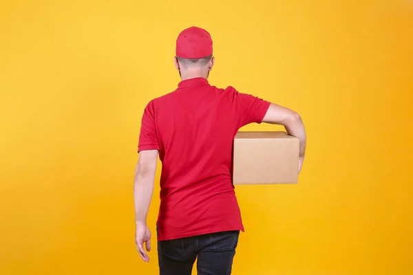 Back view of young delivery guy wearing red uniform and cap holding the blank cardboard box over isolated yellow background. Back view of bearded man carrying parcel. Copy space for text.