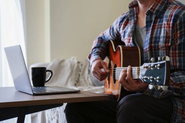Ekoseli ekose gömlekli müzik koleji öğrencisi akustik gitar egzersizi yapıyor, dizüstü bilgisayardan notlar okuyor. Karantina sırasında evde müzik dersi alan bir adam. Arkaplan, kapat