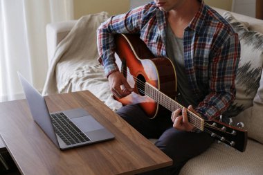 Music college hipster student in checkered plaid shirt practicing acoustic guitar exercise, reading notes from laptop computer. Man taking an online musical courses at home during quarantine. Background, close up clipart