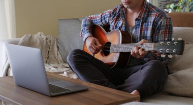 Music college hipster student in checkered plaid shirt practicing acoustic guitar exercise, reading notes from laptop computer. Man taking an online musical courses at home during quarantine. Background, close up clipart