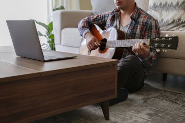 Ekoseli ekose gömlekli müzik koleji öğrencisi akustik gitar egzersizi yapıyor, dizüstü bilgisayardan notlar okuyor. Karantina sırasında evde müzik dersi alan bir adam. Arkaplan, kapat